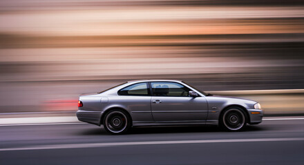 Silver luxury coupe car in motion on city street. Side view of speeding vehicle with motion blur effect. Dynamic urban transport photography. Modern automobile design concept