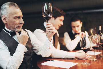 Old experienced sommelier in bow tie is holding glass of red wine while sitting at table.
