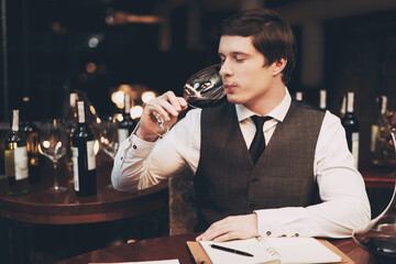  Young experienced sommelier tasting red wine, poured into glass, closing his eyes. Sommelier enjoys taste of alcoholic beverage.