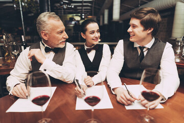 Two male sommeliers and female sommelier make up wine list sitting at table with glasses of wine. Confident sommelier tasting wine in restaurant