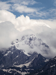 A majestic snow-covered mountain peak partially hidden by thick clouds in the Friuli Venezia Giulia region, Italy. The rugged rock formations contrast with the soft, billowing clouds.