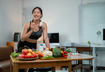 Fit asian woman wearing sportswear and headphones, enjoying nutritious meal near table with fresh produce after workout at home kitchen