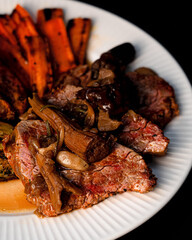 Beef tenderloin served with forest mushrooms, baked potatoes and grilled carrots on a white plate and black background. Food photography.