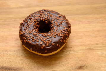 A freshly baked chocolate donut adorned with crunchy chocolate sprinkles rests on a wooden surface, showcasing its glossy glaze and inviting appearance