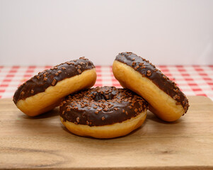 Three chocolate glazed donuts topped with sprinkles are arranged neatly on a wooden board. The background features a classic red and white checkered tablecloth