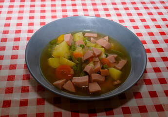 A generous serving of homemade vegetable and sausage soup is presented in a blue bowl on a vibrant red and white checkered tablecloth