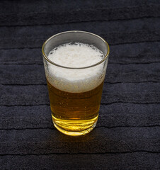 A clear glass filled with beer, exhibiting a foamy top and bubbles rising throughout. The dark background highlights the drink's vibrant color