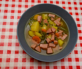 A generous serving of homemade vegetable and sausage soup is presented in a blue bowl on a vibrant red and white checkered tablecloth