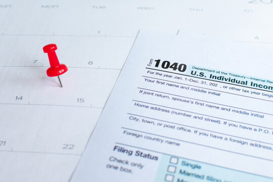 Close up shot of a red drawing pin (thumbtack or pushpin) on a calendar on the 15th. With Form 1040 U.S. Individual Income Tax Return. Selective focus. Tax due date, taxation reminder, extension