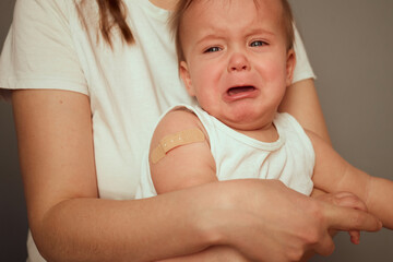 A young child in a white shirt is held by a parent after receiving a vaccination. The child shows signs of distress, indicating discomfort from the recent inoculation on the arm