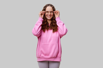 A young woman stands with a playful expression, adjusting her glasses while wearing a cozy pink hoodie. Her long hair is styled in waves, creating a warm and inviting atmosphere