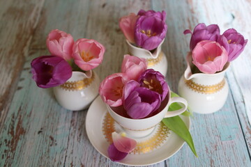On the wooden table, there is a cup filled with purple tulip flowers and three milk jugs with...