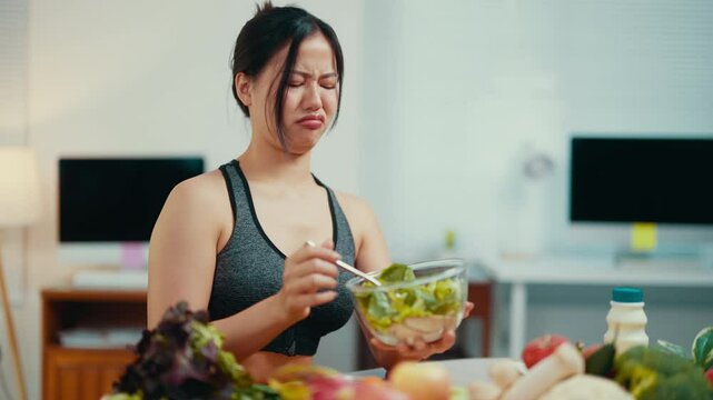 Young asian sportswoman grimacing, sitting at table with fresh produce, experiencing strong distaste while eating salad, demonstrating nutrition resistance