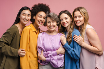 Five multiethnic women of different ages hugging and smiling on pink background