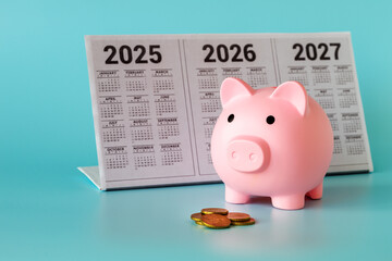 A piggy bank in the form of a pink pig, a desktop calendar for 2025-2027 and a pile of coins on a light blue background. The concept of long-term investments.