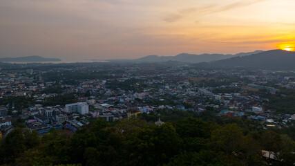 Fototapeta premium A scenic view of a town nestled among lush green mountains at sunset, with a temple structure perched on the hillside and a vibrant orange sky creating a warm, tranquil atmosphere.