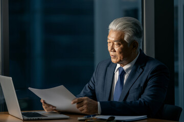 A man in a suit is sitting at a desk with a laptop and a stack of papers