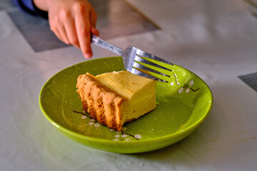 A piece of cheesecake on a green plate, from which a piece is being broken off with a fork.