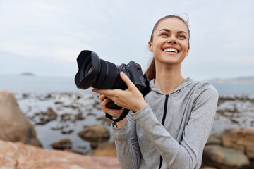 Smiling young woman in sportswear holding a camera by the seaside, capturing nature s beauty, showcasing joy and creativity in photography