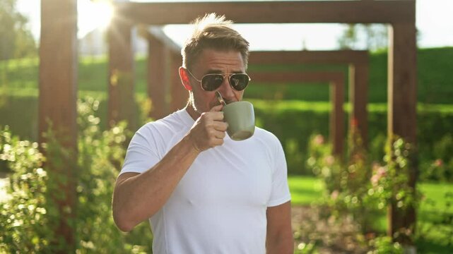 Mature man wearing sunglasses drinking cup of coffee in home garden at morning. Handsome middle aged male in 50s standing under wooden pergola on terrace. Backlit by sun, lensflare.