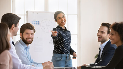 Young Indian woman standing at front of participants, pointing toward staff member, ask question, prompts response, fosters interaction, encourages involvement, engaged in positive lively discussion