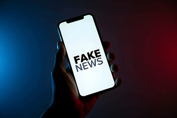 A hand holding a smartphone displays the text "fake news" against a dark background with blue and red lighting, symbolizing the impact of misinformation in media