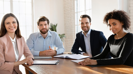 Four business partners take part in meeting gathered at conference table, smile, look at camera engaged in virtual videocall negotiations with shareholders for exchanging information. Staff portrait