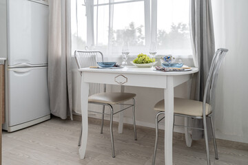 a simple, cozy kitchen in bright colors, a romantic breakfast on a white kitchen table by a large window. There are plates, glasses and grapes on the table.