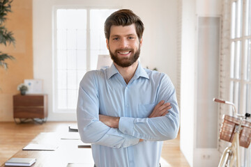 Fototapeta premium Portrait of young businessman, coach or business trainer standing confidently in modern sunny conference room, smiling, looking at camera, reflecting his success or aspirations in professional field