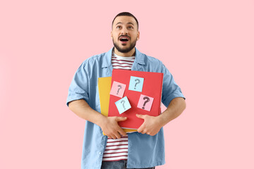 Portrait of surprised young man holding folders with question marks on pink background
