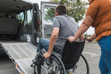 Female assistant pushing a man with disability, a wheelchair user up the car ramp into the van. Accessibility and travel concepts.
