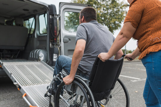 Female assistant pushing a man with disability, a wheelchair user up the car ramp into the van. Accessibility and travel concepts.