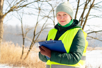 Female ecologist inspects trees in winter. Middle aged female volunteer with a tablet checks trees in the forest after a snowfall. Forestry engineer works in winter. Inspector monitors forest lands.
