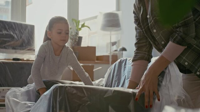 Little girl and her mother covering armchair with protective cellophane while bending over furniture and preparing for renovation of their new apartment