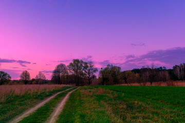 View of the countryside with dirt road and dramatic purple evening sky during sunset. Rural landscape in spring