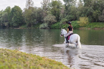 Girl riding a snow white horse down the calm river water with forest greenery reflections from the...