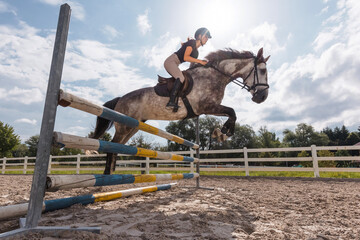 Girl rider on a dapple gray horse jumping over a hurdle at the manege of equestrian center on a...