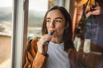 Young relax woman smoking electronic cigar near window at home