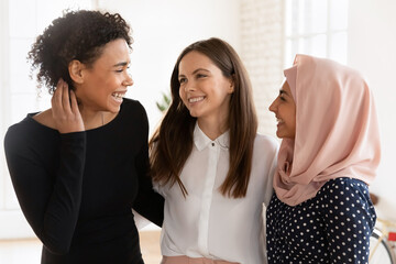 Multicultural women colleagues, students, friends hugging standing together indoor, enjoy effective teamwork, result and reached goals, having warm relationships at work. Friendship, equality, loyalty
