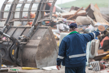 Fototapeta premium Landfill worker directing skid steer loader on the garbage heap, rear view. Waste disposal, consolidation, and transfer concept.