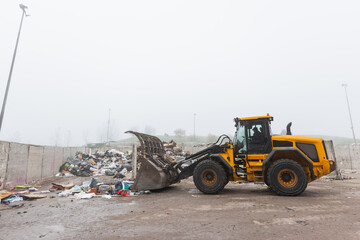 Yellow wheel loader, with lifted scrap grapple, moving along the recycling center area in process handling dumped waste