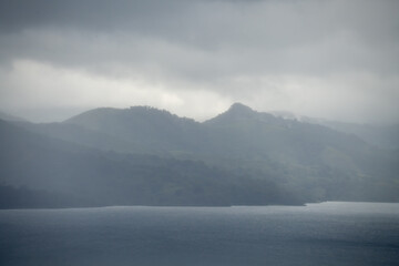 Misty mountains rise from the edge of a calm lake under a cloudy sky..