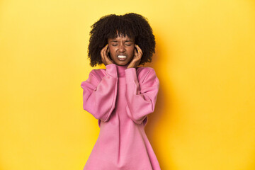 Teen girl in pink sweatshirt, yellow studio backdrop covering ears with hands.