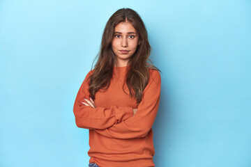 Teen girl in vibrant orange sweater on blue suspicious, uncertain, examining you.