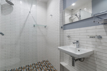 The bathroom is decorated with white tiles. A mirror over a small sink, a shower with a tropical watering can behind a glass partition.