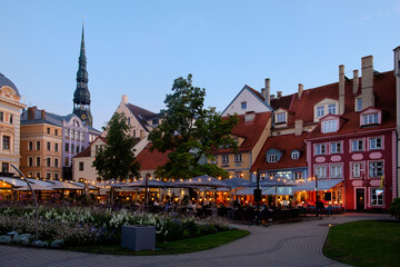 Obraz premium A scenic view of Livu Square in Old Town of Riga, Latvia, featuring colorful historic buildings, lush greenery, and outdoor cafes in a popular tourist destination.