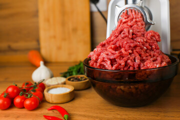 Side view on a wooden background: dark dishes with red minced meat inside, which comes out of a metal meat grinder, spices, herbs and vegetables are nearby. The concept of healthy eating, cooking