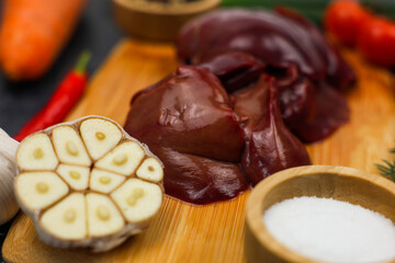 Close-up of raw chicken liver on a wooden plank surrounded by vegetables, seasonings in wooden bowls and herbs on a dark background. The concept of healthy eating, cooking, pate recipe. High quality