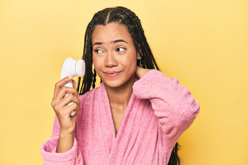 Indonesian woman with facial cleaner on yellow touching back of head, thinking and making a choice.