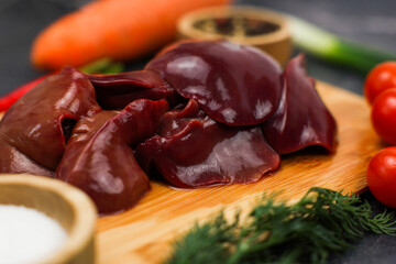 A close-up of poultry liver in the center lies on a wooden cutting board, surrounded by vegetables, spices in wooden bowls, and herbs. The concept of proper and healthy nutrition, vitamins and trace